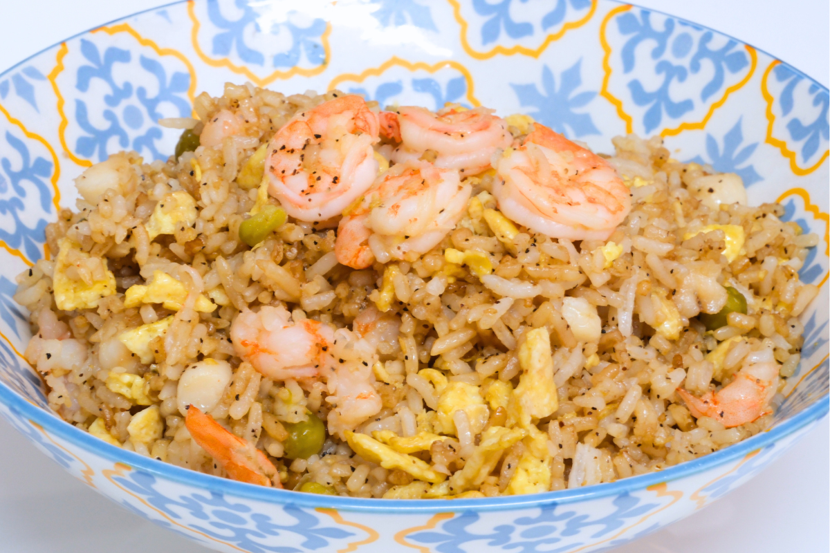 A bowl of golden Mixed Seafood Fried Rice with shrimp, fish, eggs, and green peas, served with a side of soy sauce.