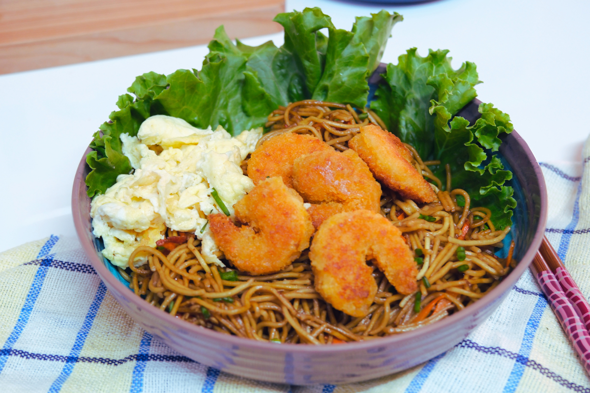 A plate of stir-fried egg noodles tossed in dark soy sauce, topped with crispy Fishta Popcorn Shrimp, scrambled egg, and julienned carrots, served hot in a stylish bowl.